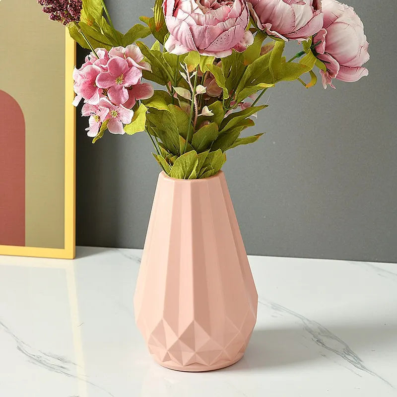 A pink faceted ceramic vase filled with pink peony artificial flowers and green leaves, set on a white marble surface against a gray wall.