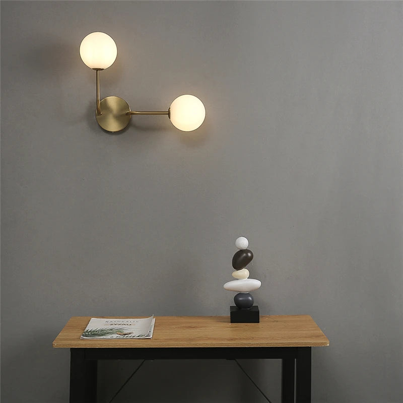 Wall-mounted lamp with two round lights on a gray wall above a wooden table with a magazine and small sculpture.