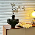 A ribbed black ceramic vase with white blossoms in a dark branch, styled on a wooden side table next to an open book and a glowing table lamp in front of white window blinds.