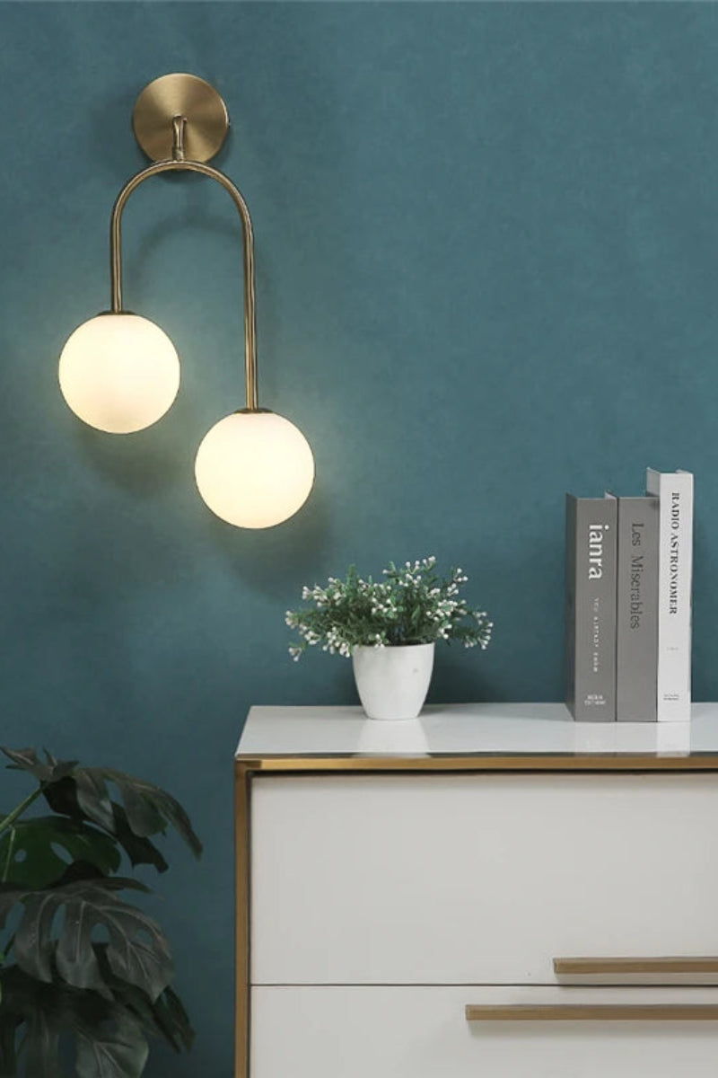 Wall sconce with two round lights on a teal wall above a white dresser with books and a plant.