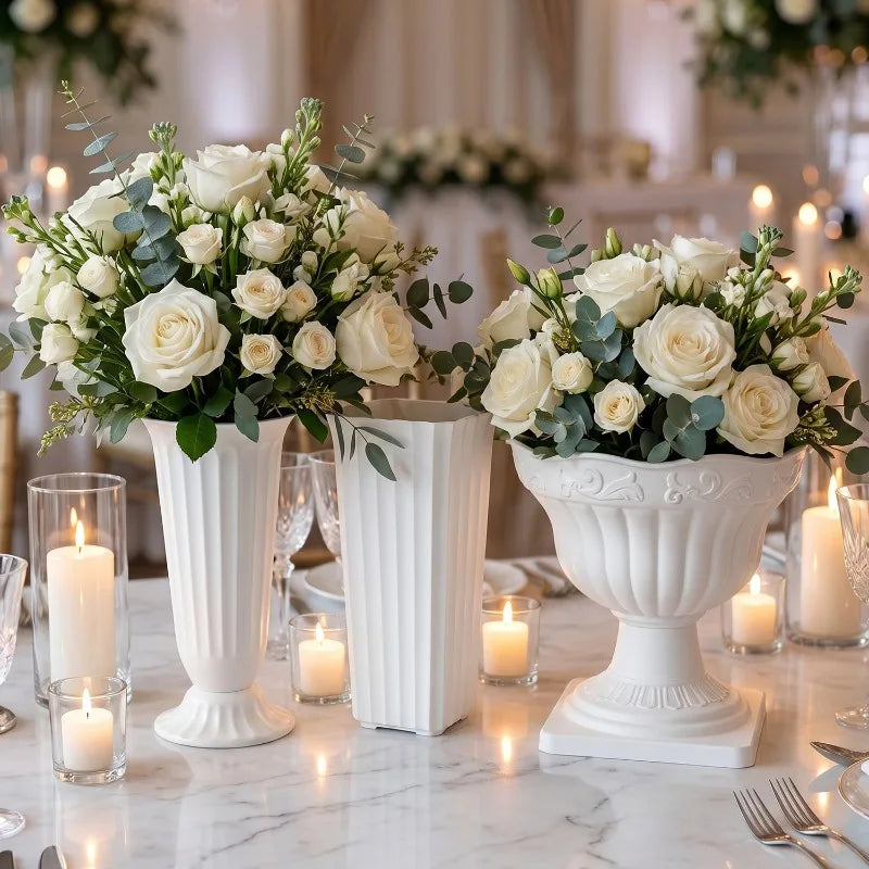 Three white floral centerpieces with white roses, green foliage, and eucalyptus stems displayed in white pedestal vases on a marble table with lit candles.