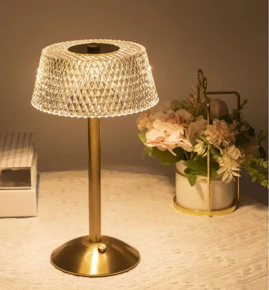 A light luxury crystal desk lamp with a gold-finished metal base and a textured, faceted clear crystal shade, shown illuminated on a tabletop next to flowers and a book. 
