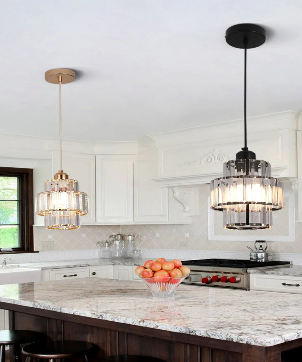 Two modern pendant lights hanging in a bright white kitchen with dark wood cabinetry and a marble island. The right pendant has a black metal frame and clear glass shades, while the left pendant has a gold-toned frame and clear glass shades.