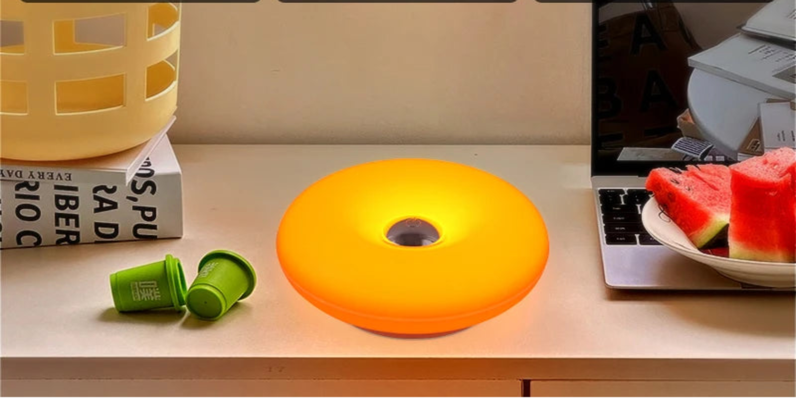 A color-changing LED ring light shown on a desk with three panels above demonstrating warm light (3000K), neutral light (4500K), and white light (6000K). The ring light itself is glowing orange/yellow in the foreground, with books, a laptop, and watermelon on the desk.