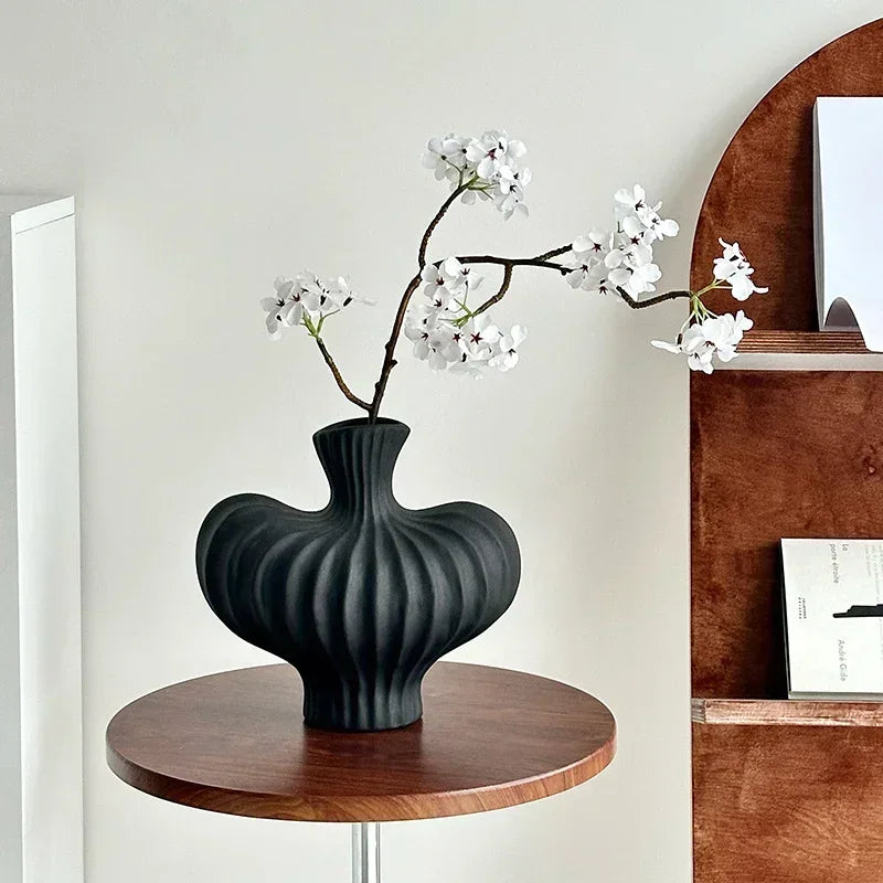A ribbed matte black ceramic vase with a flared neck, holding a flowering branch with white blossoms, displayed on a round wooden side table in a modern interior setting.
